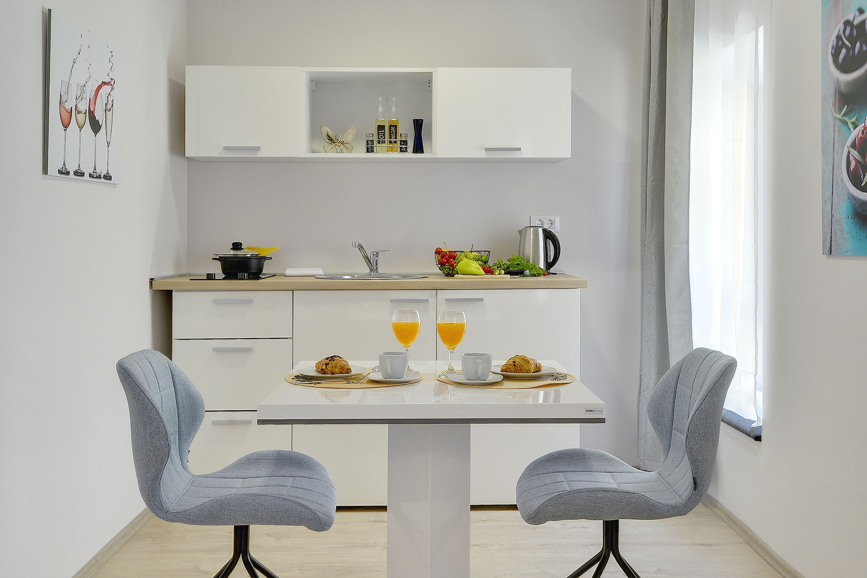 A modern kitchen with white cabinets, a dining table with two chairs, and a breakfast setup including croissants, juice, and a coffee pot.