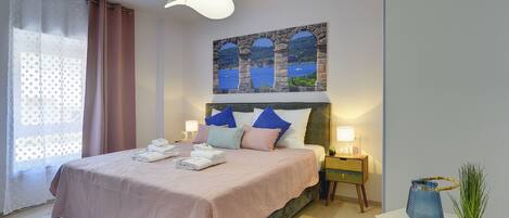 Una cama bien hecha con colcha rosa y almohadas azules, un cuadro de una escena costera sobre ella y una mesita de noche con una planta y una lámpara al lado.