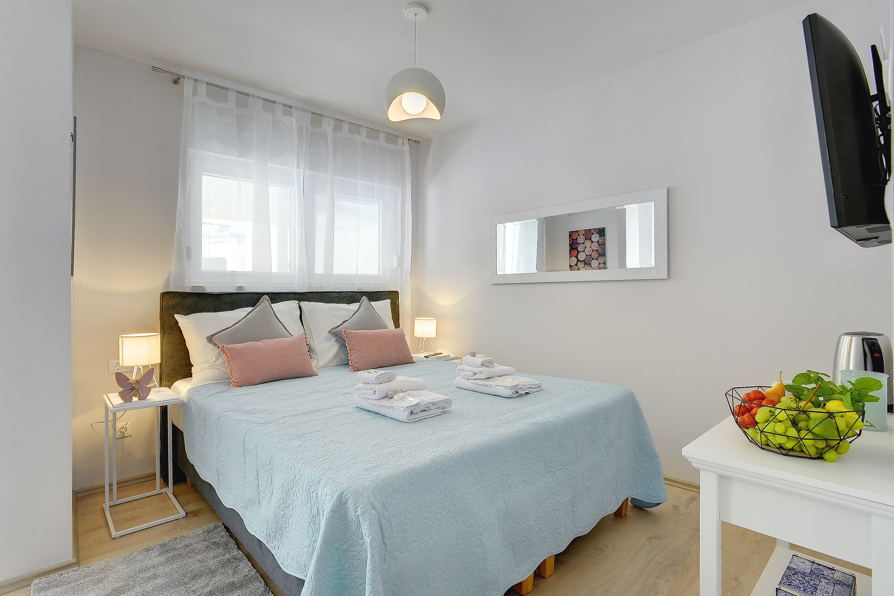 A neatly made bed with a light blue bedspread, a bedside table with a lamp, and a fruit bowl on a side table.