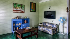 A room with a wooden table, a blue cabinet, a patterned sofa, a television, and a standing fan. - Casa Cantinho em Ilhabela (Ilhabela)