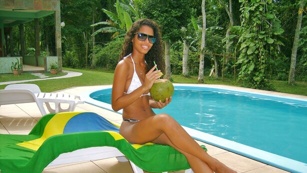 Una persona sentada en una silla de descanso junto a una piscina, sosteniendo un coco.