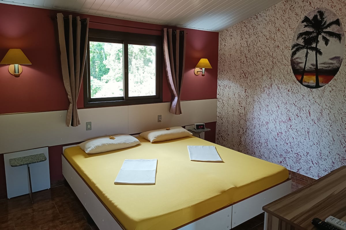 Una habitación con una cama, dos lámparas de pared, un ventanal con cortinas, un reloj de pared y un mural tropical.