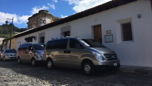 A row of parked vans in front of a white building with a tiled roof, a sign with the number 19E, and a small window with a sign. - Suite El Nido (Antigua Guatemala)