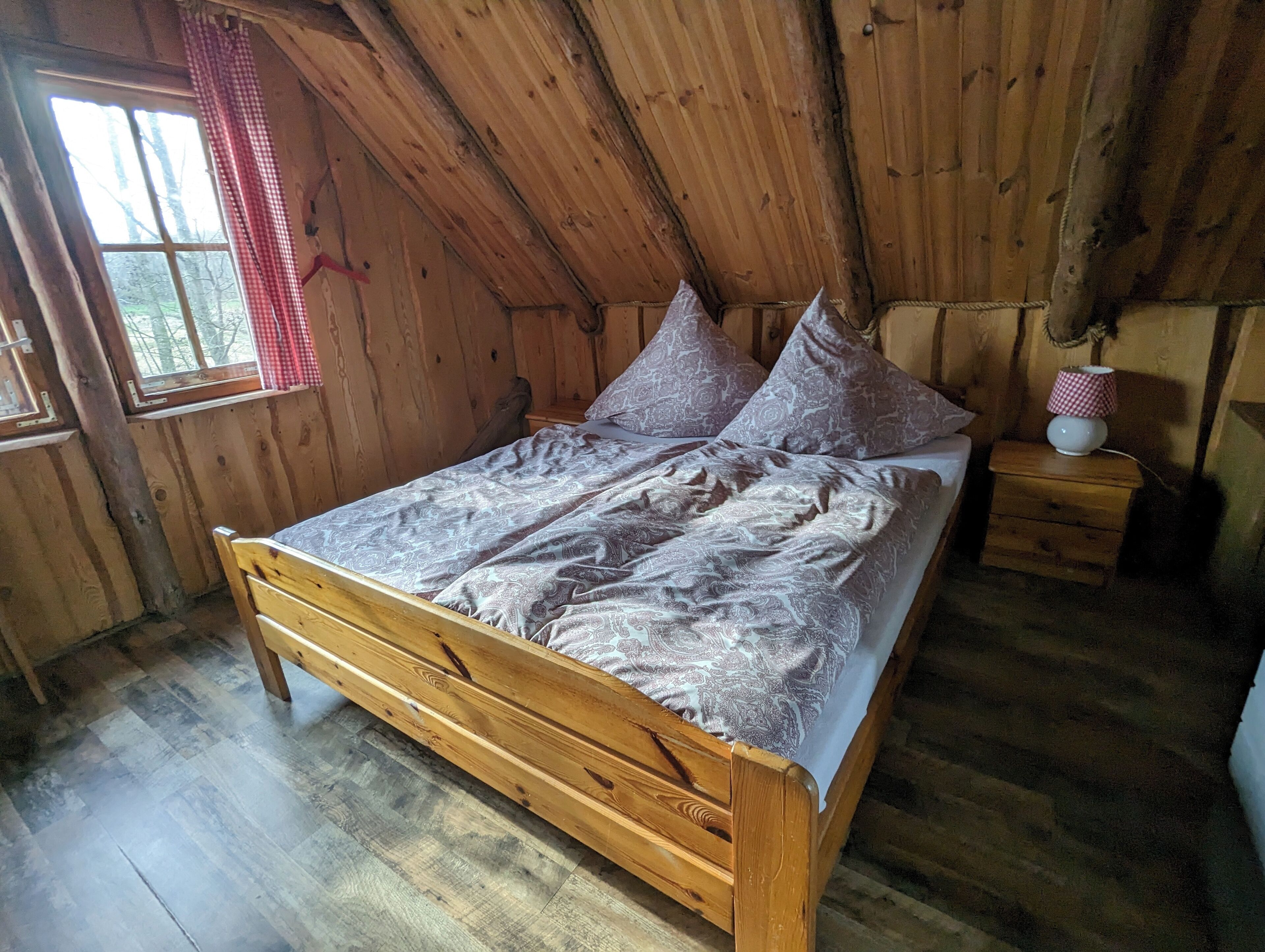 Ein hölzernes Bett in einem Zimmer mit schrägem Dach, ein Fenster mit roten Karoo-Vorhang und ein Nachttisch mit Lampe.