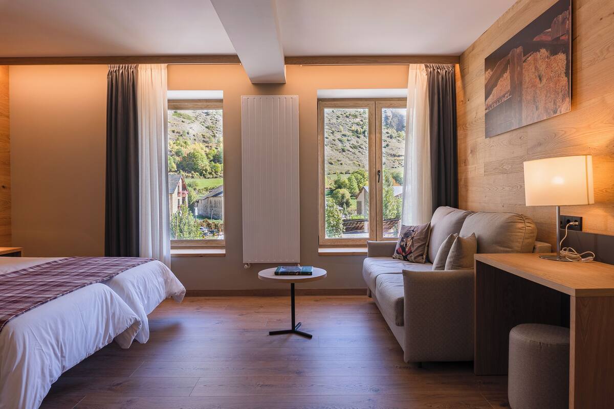 A hotel room with a bed, a sofa, a small table, and a window with curtains.