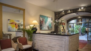 Un área de recepción con mostrador de mármol, sillas de mimbre, un espejo grande, pinturas abstractas y vistas del patio exterior.