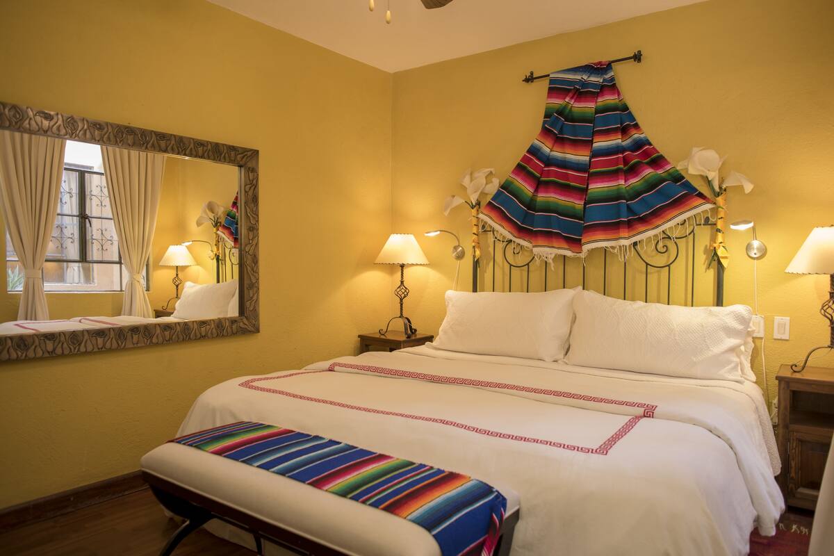 Une chambre d’hôtel comprenant un lit, une banquette, un miroir, une fenêtre avec des rideaux et une lampe fixée au mur.