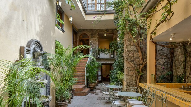 Un patio con una fuente, plantas en macetas, mobiliario exterior y una escalera que lleva a un nivel superior.
