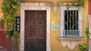 Un edificio con un gran puerta doble de madera, una ventana con barrotes de metal y flores rojas, y un letrero que dice "CASA".