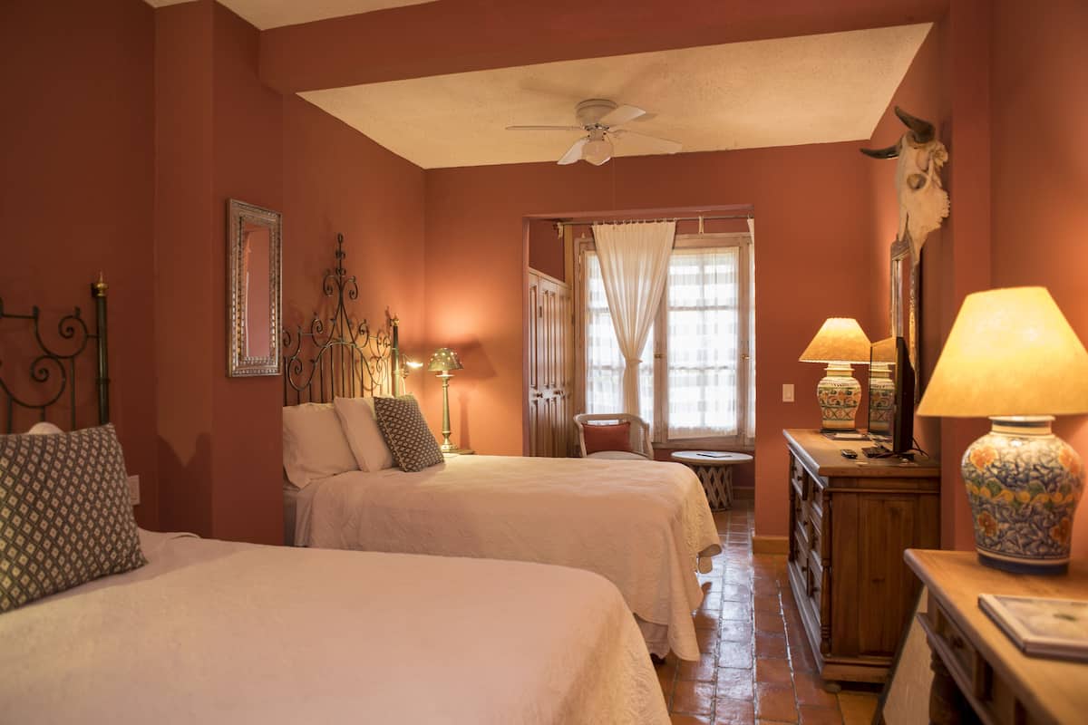 Une chambre avec deux lits, un ventilateur de plafond, une fenêtre avec des rideaux, une lampe et un vase décoratif.