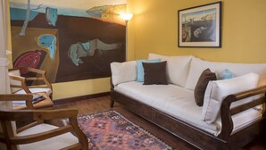 Una sala de estar con un sofá blanco, sillas de madera, un tapete estampado y dos cuadros en las paredes.
