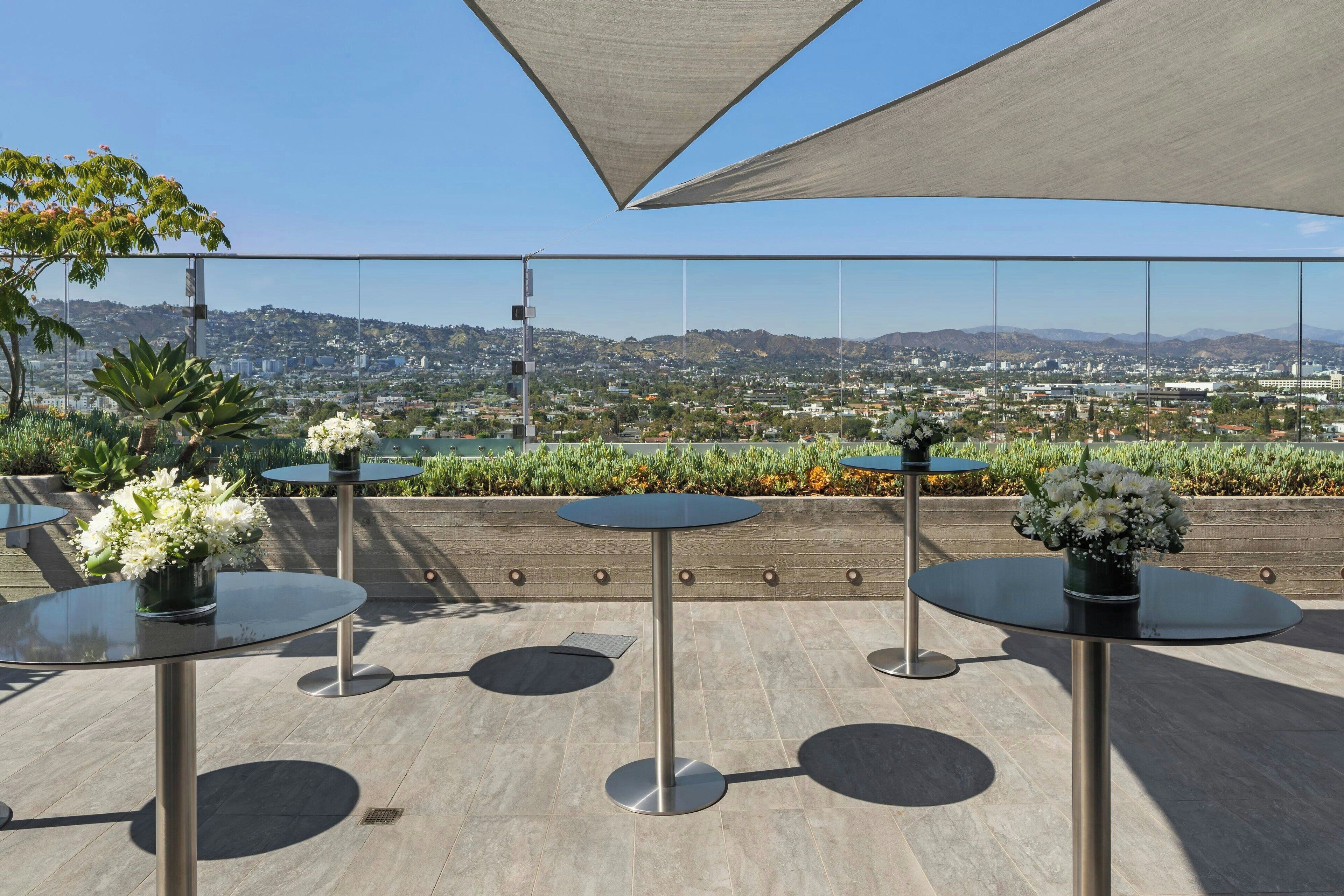 A rooftop terrace with round tables, potted plants, and a view of a cityscape.