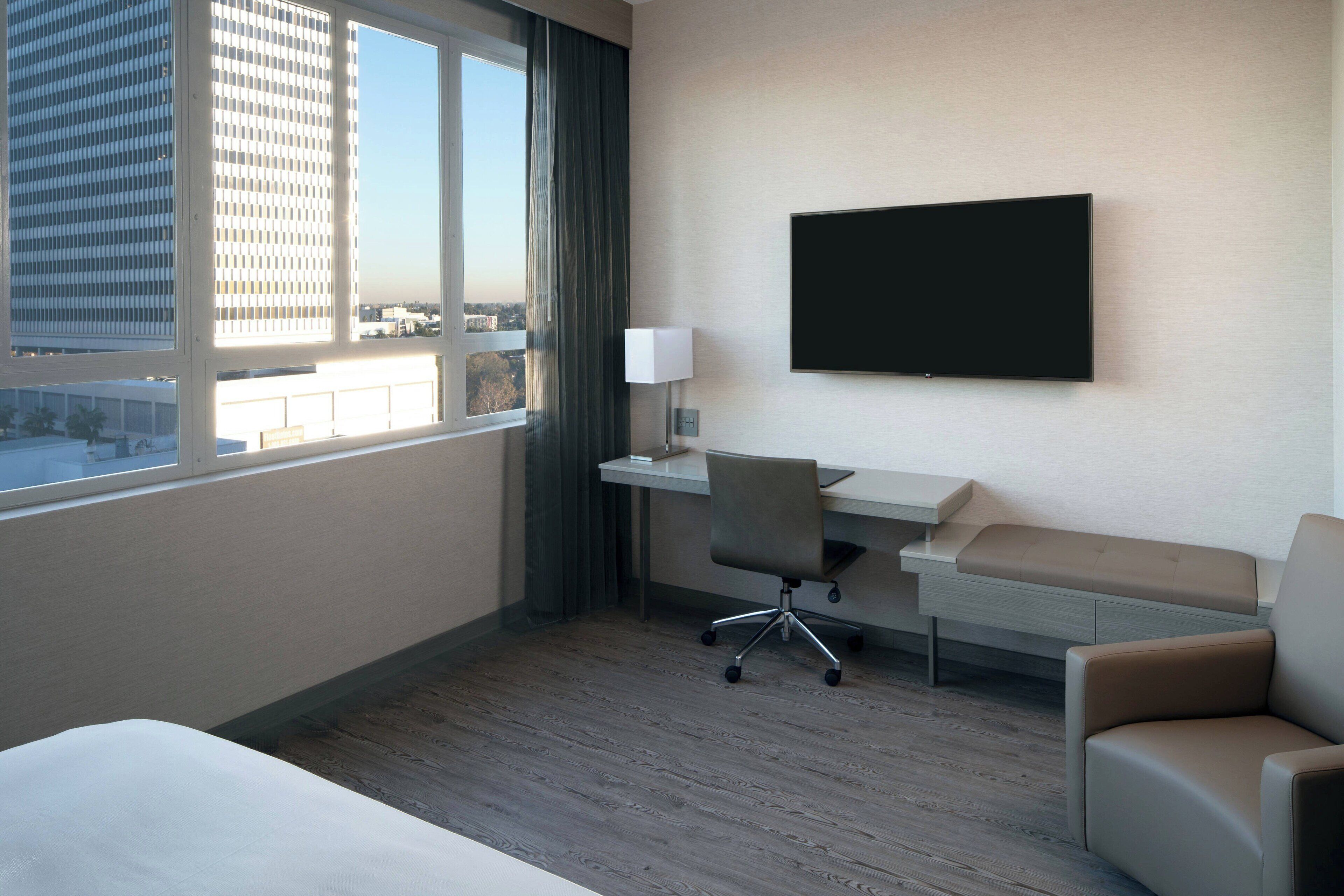 Une chambre d’hôtel comprenant un lit, un bureau avec une chaise, une télévision et une vue sur le paysage urbain, visible à travers une grande fenêtre.
