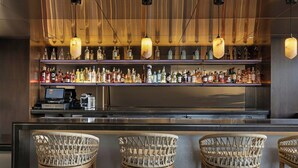 Een bar met een lange toonbank, meerdere flessenkasten en drie rieten barkrukken.