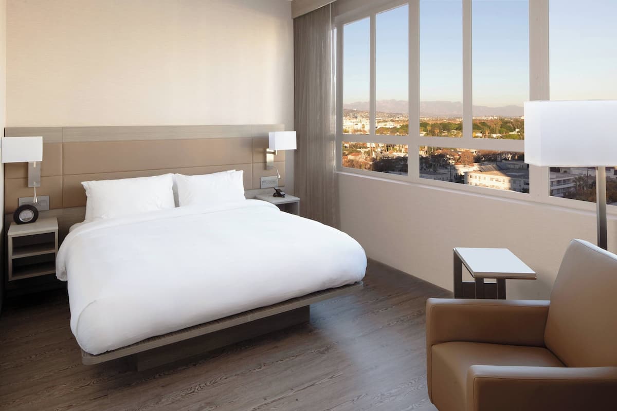 Une chambre d’hôtel avec un grand lit, des tables de chevet, une chaise et une vue sur le paysage urbain, visible à travers une grande fenêtre.