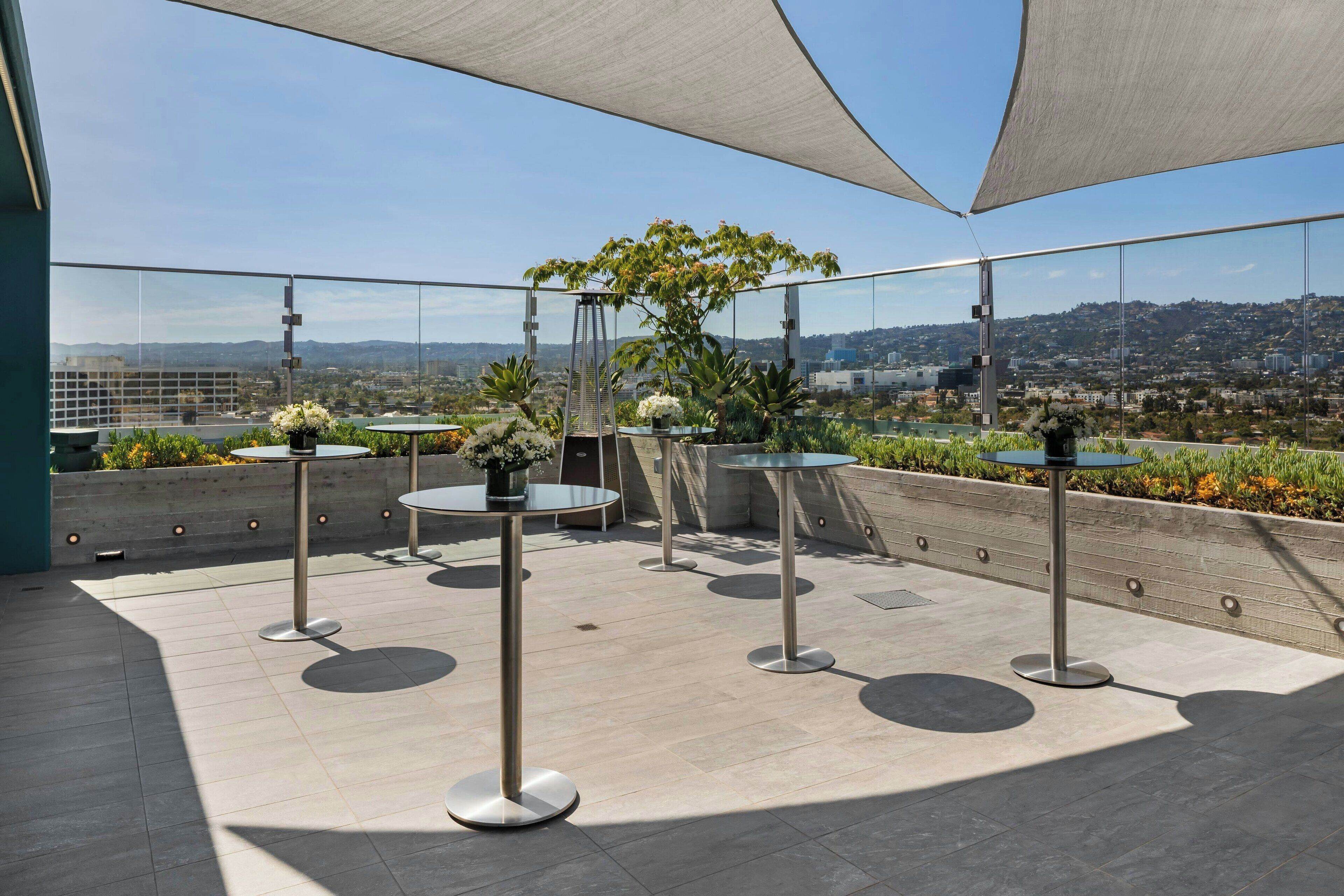 A rooftop terrace with round tables, potted plants, and a clear view of the cityscape.