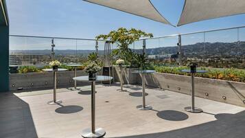 A rooftop terrace with round tables, potted plants, and a clear view of the cityscape.