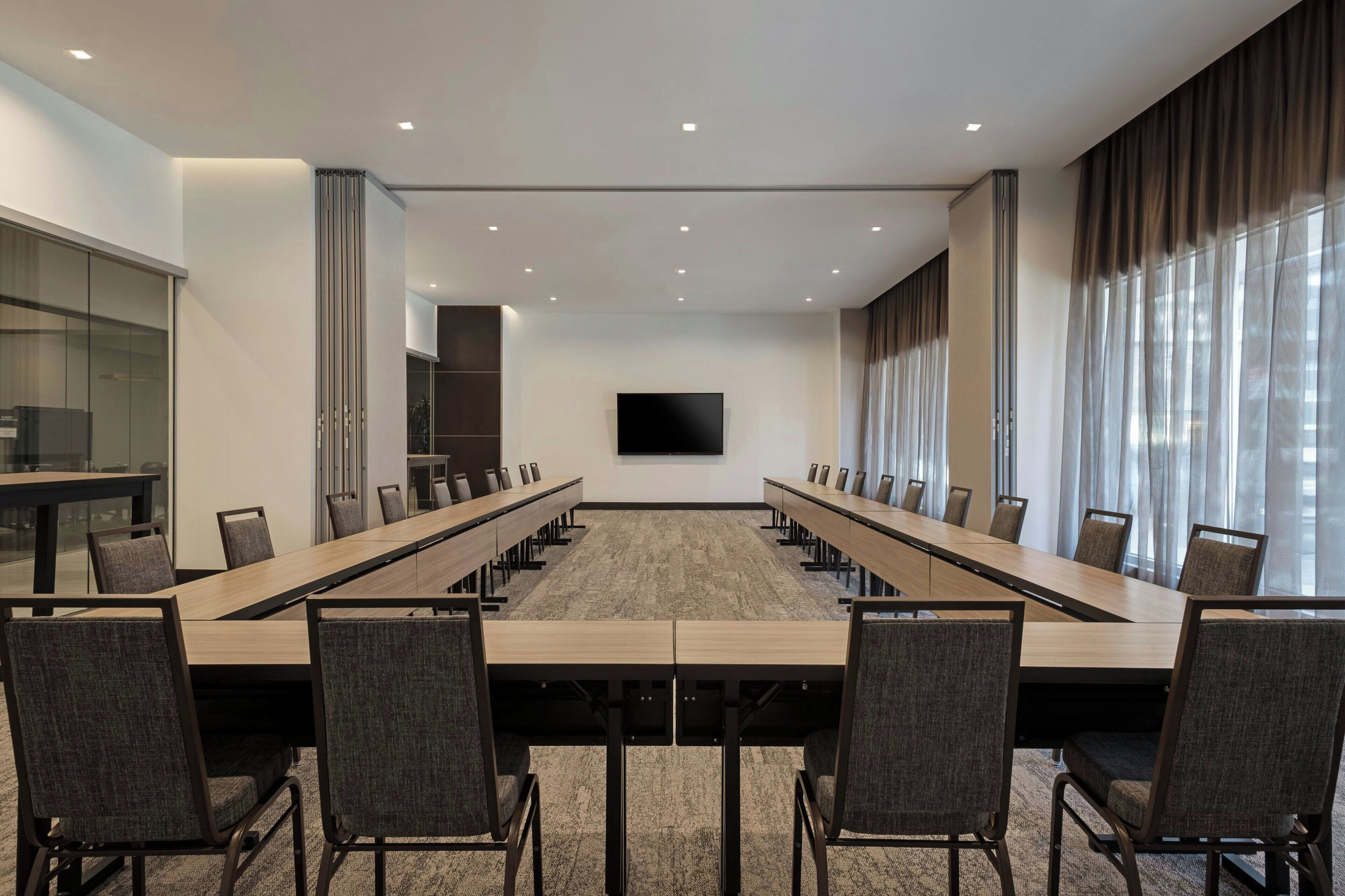 A conference room with a long table, chairs, a flat-screen TV, and large windows with curtains.