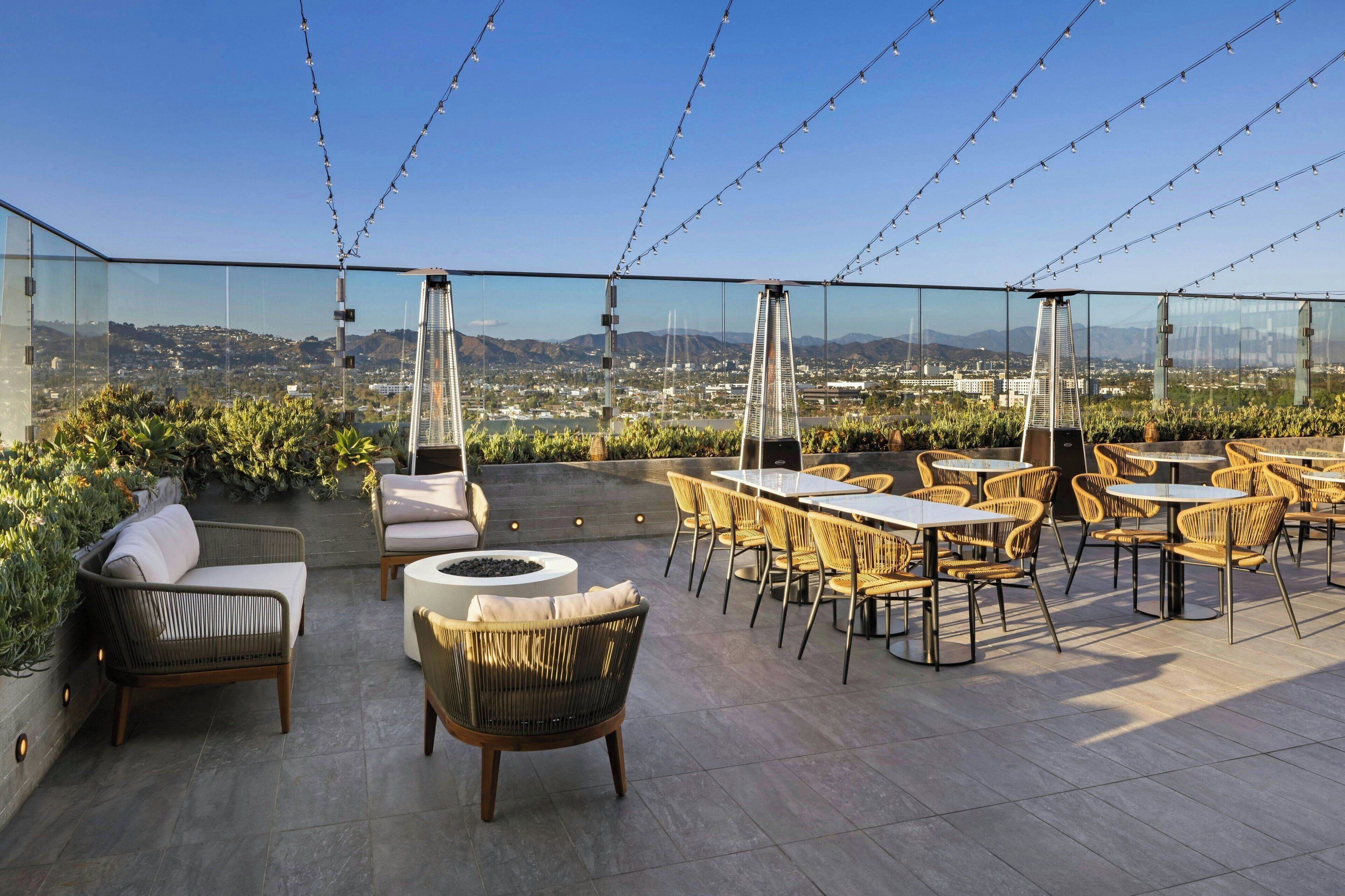 An outdoor terrace with wicker furniture, a fire pit, and a glass wall offering a city view.