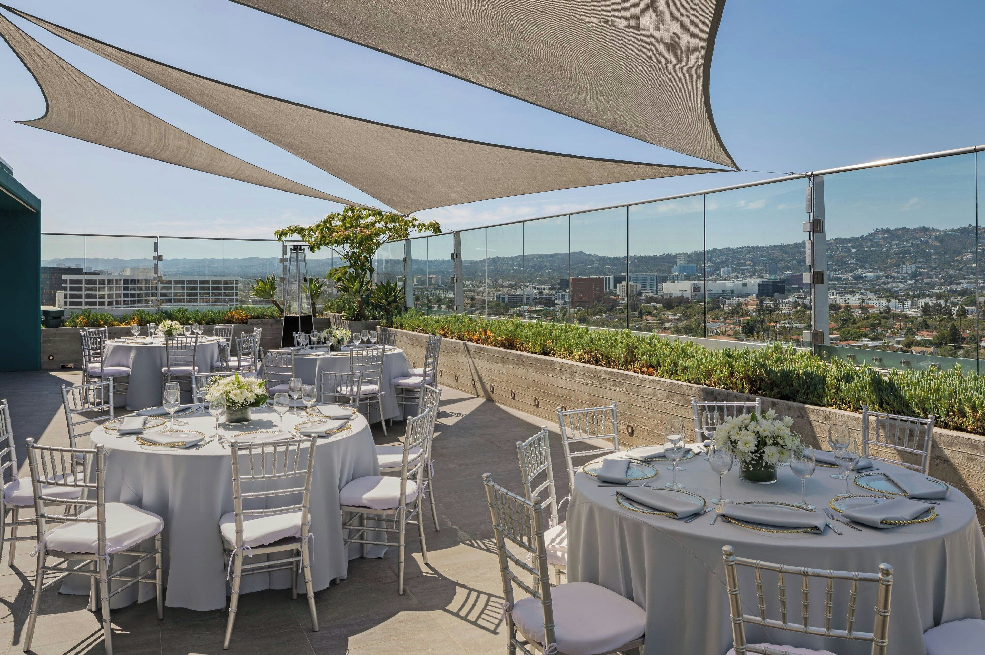 An outdoor dining area with round tables set for a meal, surrounded by white chairs, under large shade sails.