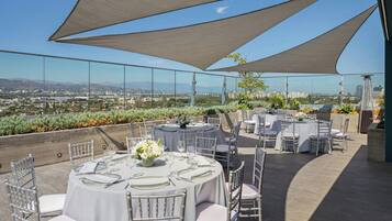 Area pranzo all'aperto con tavoli rotondi e sedie bianche, con vista sullo skyline della città.