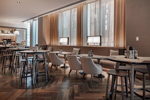 Un comedor moderno con varias mesas y sillas, ventanas amplias con cortinas translúcidas y un suelo de madera con diseño en espiga.
