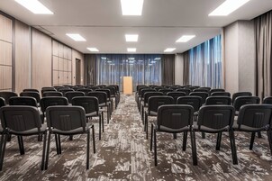 Un salón de conferencias con filas de sillas, un atril y ventanas grandes con cortinas.