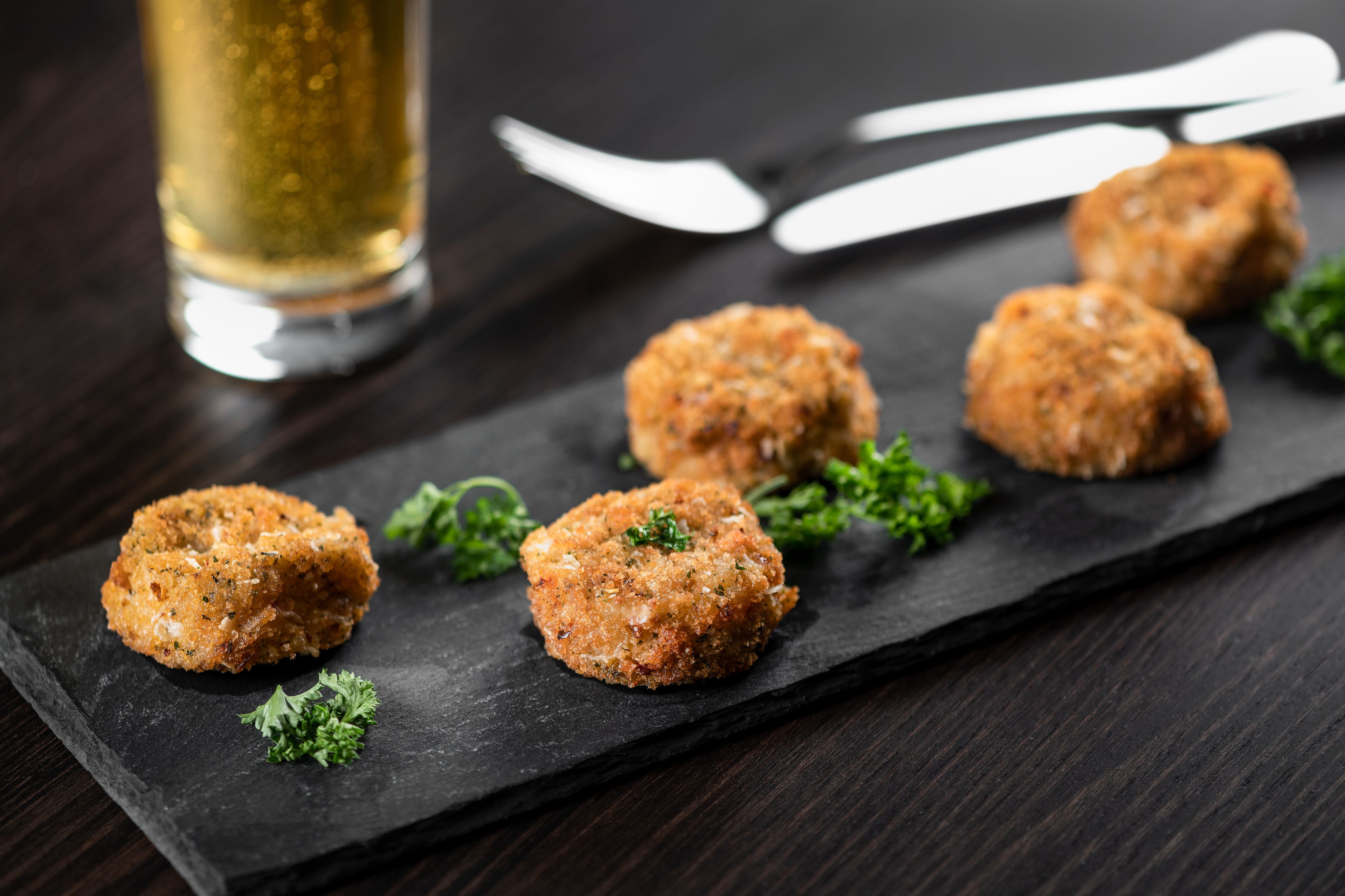 A slate serving board with five round, breaded items garnished with parsley, accompanied by a glass of golden liquid and cutlery on a dark wooden table.