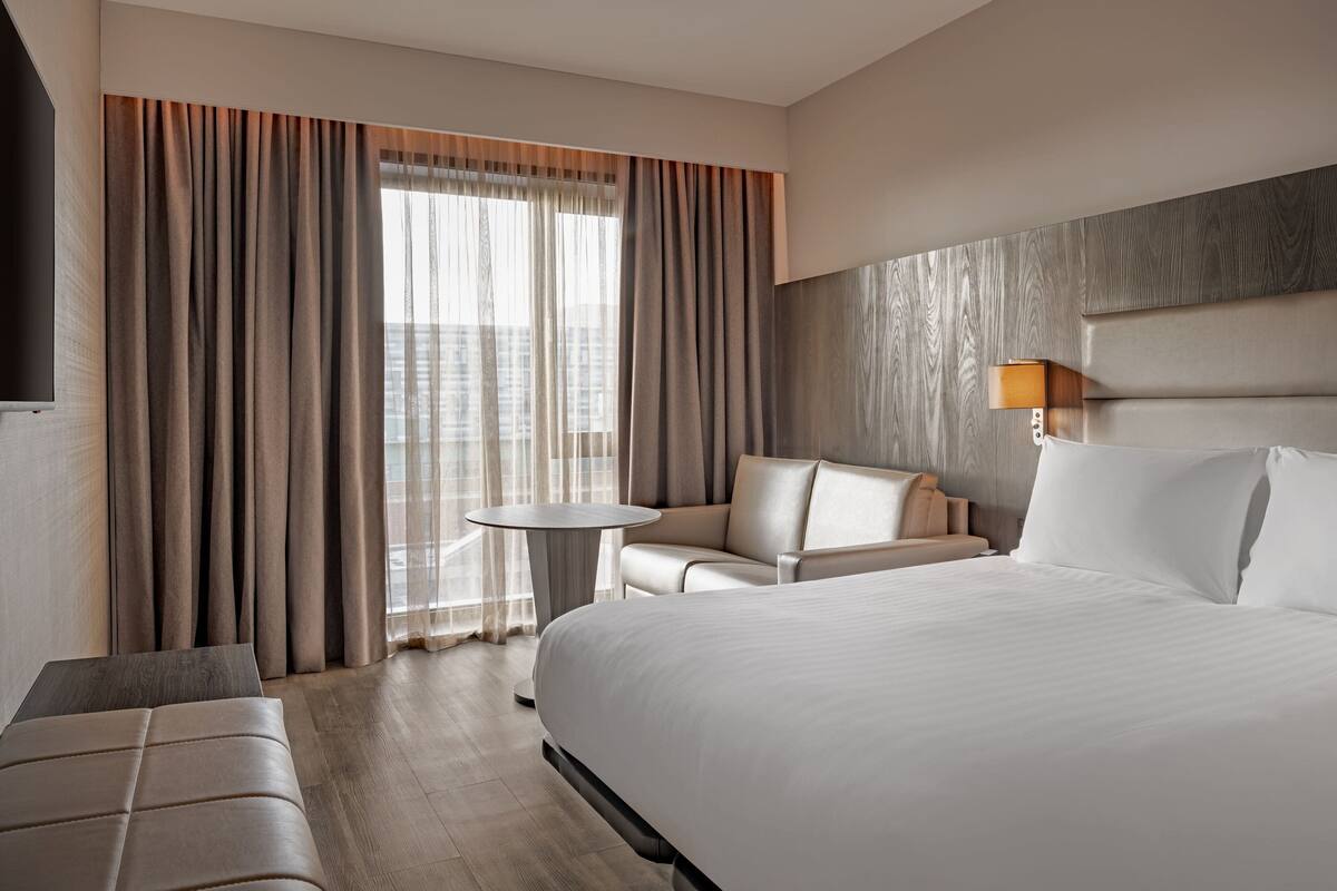 Ein Hotelzimmer mit einem großen Bett, einer Sitzecke mit Tisch und Blick auf die Stadt durch große Fenster.