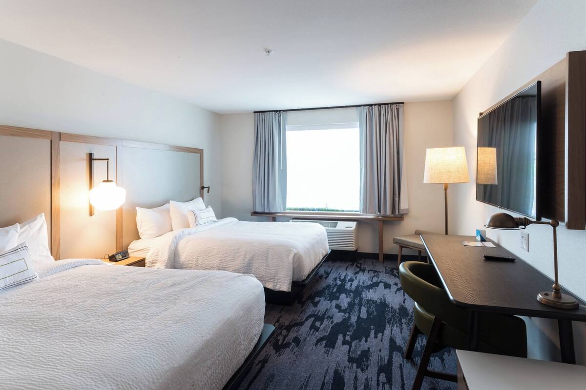 A hotel room with two beds, a desk, a chair, a lamp, a large window with curtains, and a flat-screen TV.
