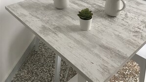 Une table en bois rustique, sur laquelle reposent deux pichets blancs, une petite plante en pot et une théière noire, le tout posé sur une nappe blanche.