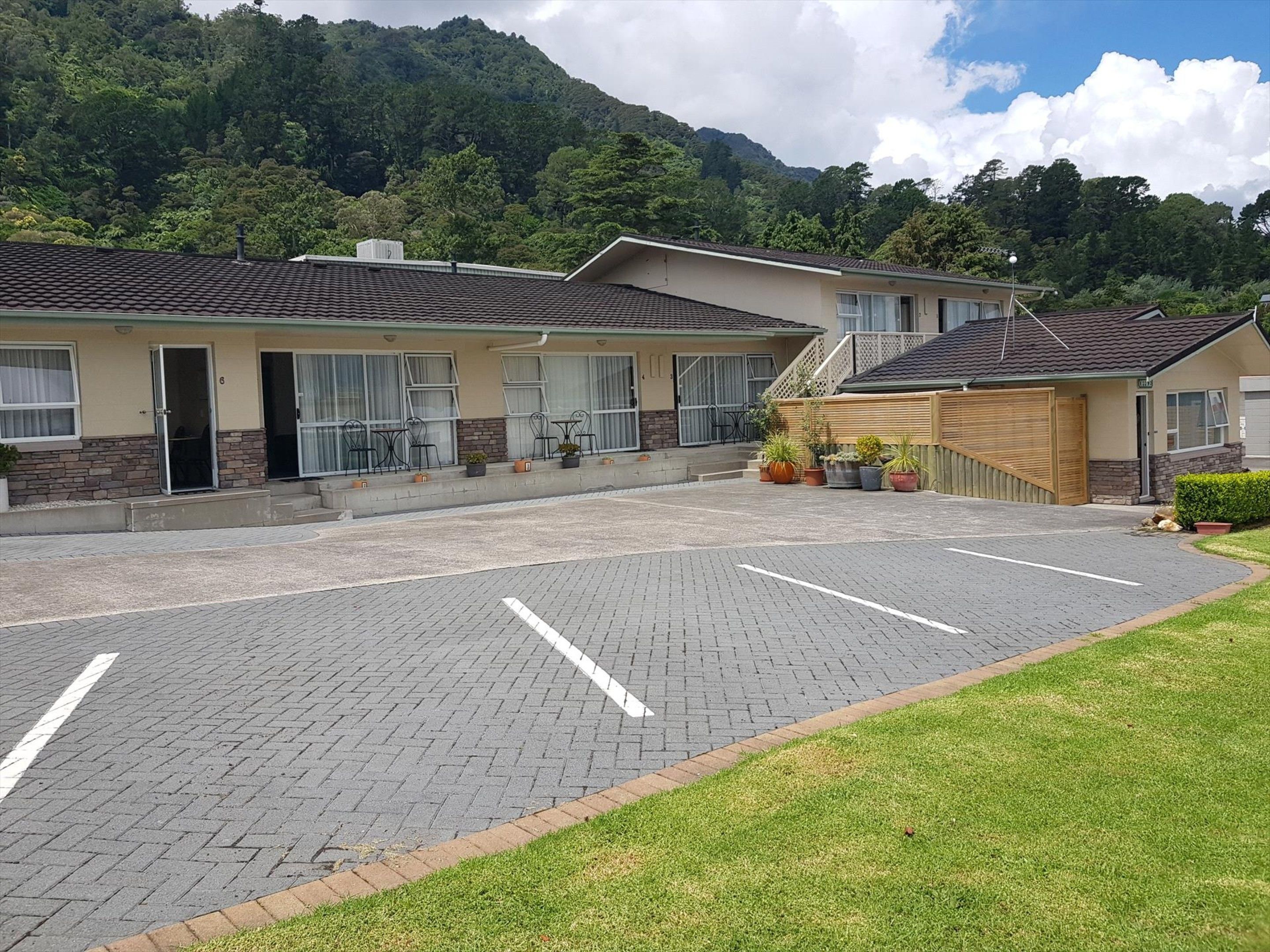 A hotel with a paved parking area, a covered outdoor seating area, and a mountain backdrop.