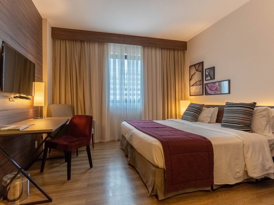 Habitació d'hotel amb un llit, una taula de treball amb cadira, una televisió penjada a la paret i una finestra amb cortines.