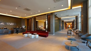 Een hotel lobby met een receptiebalie, een rode bank en een zithoek met ronde tafels.