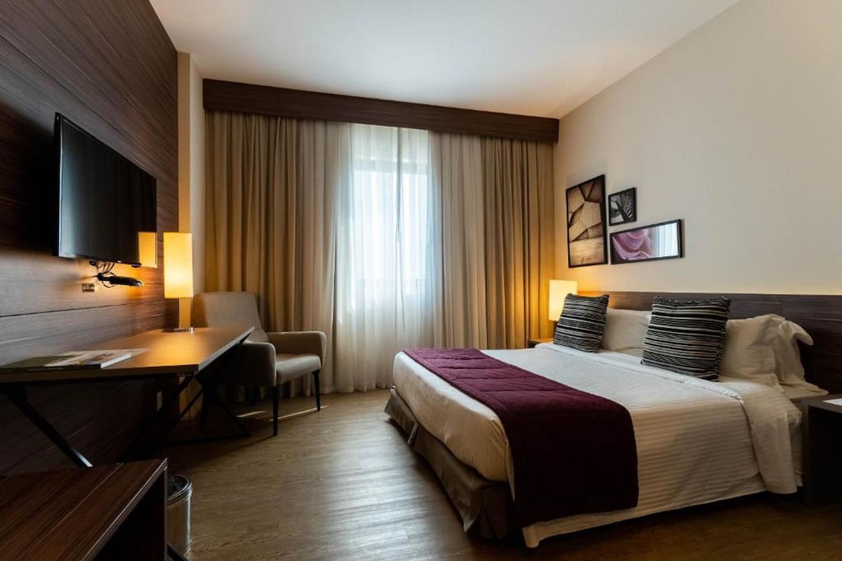 A hotel room with a bed, a desk, a chair, a TV, and a window with curtains.