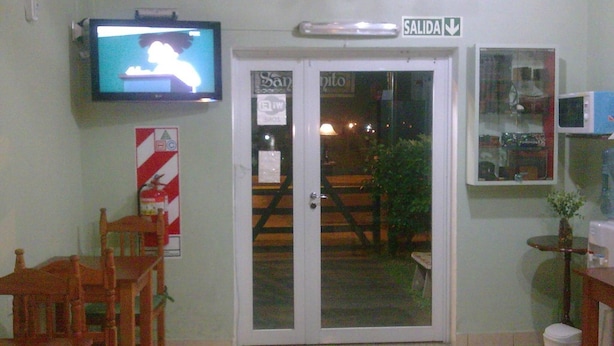 Una habitación con una televisión montada en la pared, una puerta con el letrero 'SALIDA', una silla, una mesita y un microondas.
