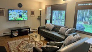 A living room with a leather sofa, a glass coffee table, a flat-screen TV, and a large window with a view of trees. - Your Cozy Anchorage Retreat (Anchorage)