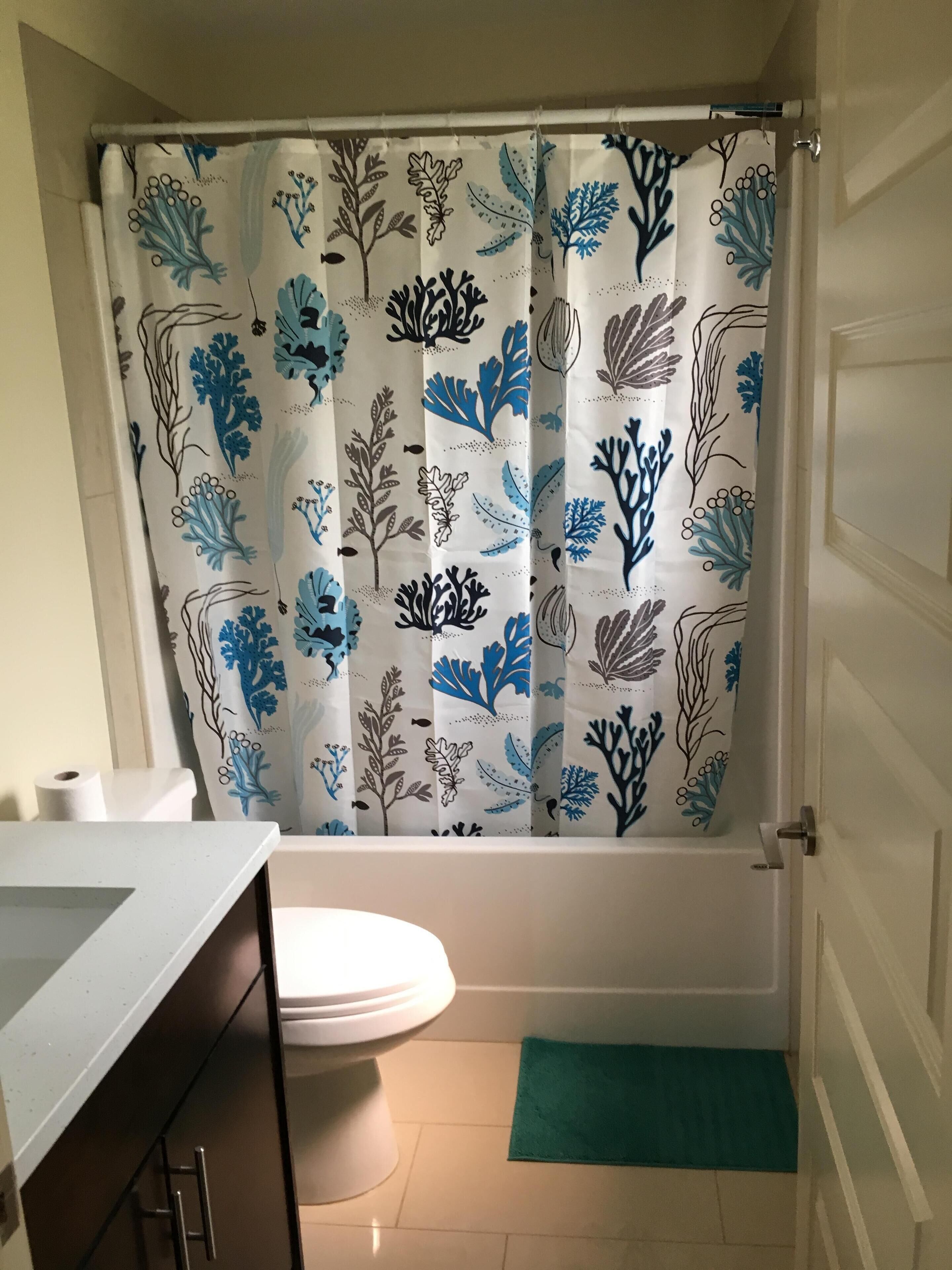 Un baño con cortina de ducha con estampado floral, inodoro, lavamanos y un tapete verde en el suelo.