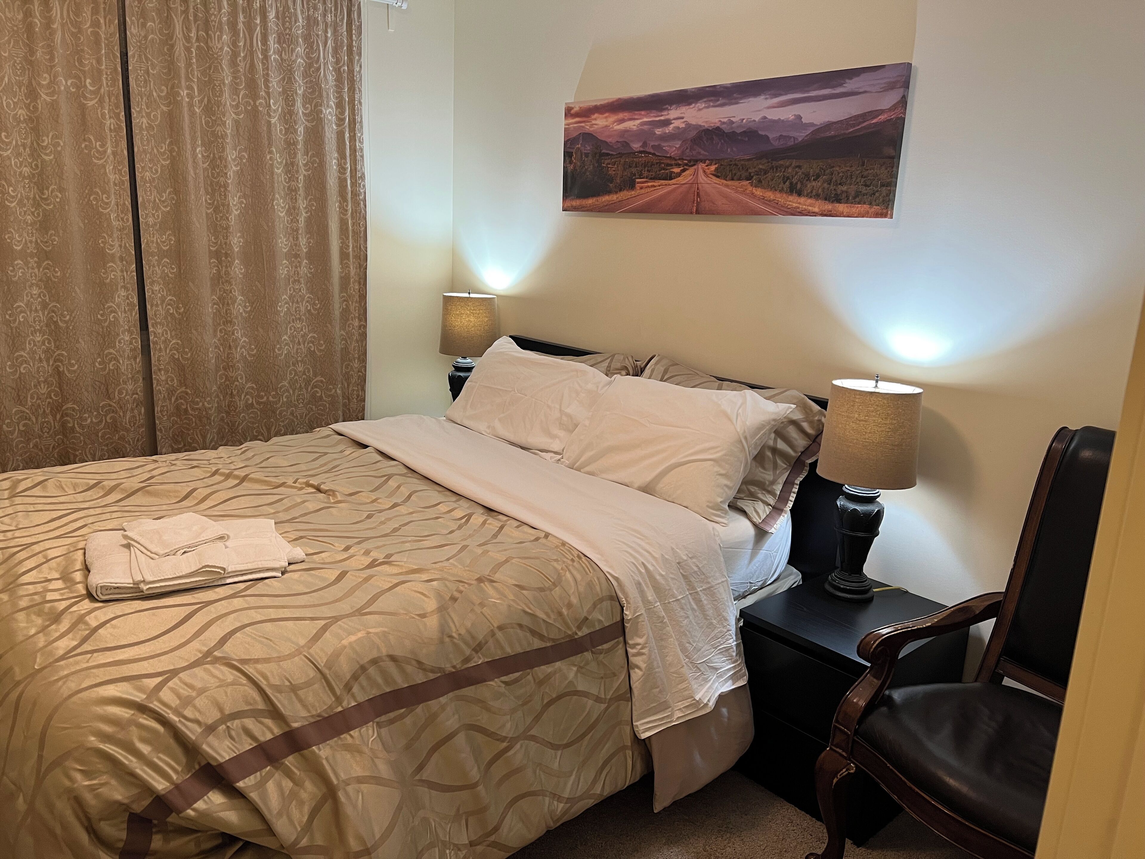 Een net opgemaakt bed met een bedsprei met patroon, een opgevouwen handdoek op het bed, een lamp op een nachtkastje en een stoel in de hoek.