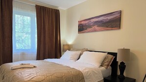 A neatly made bed with a beige patterned bedspread, a bedside lamp, and a painting of a landscape on the wall. - Modern Vacation Home Near West Edmonton Mall, Transit Center & LRT - 3BR (Edmonton)