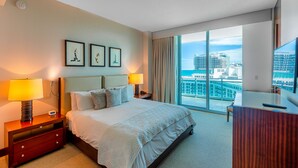 A hotel room with a large bed, bedside lamps, a desk, and a view of the city through large windows. - One Bal Harbour Hotel and Resort, Deluxe 2 bedroom private unit @ the Ritz (Bal Harbour)