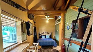 Una habitación compacta con cama, una pequeña mesa con taburetes, ventilador de techo y vista al exterior.