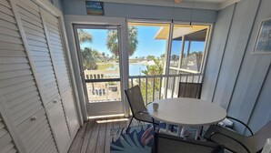 Une pièce avec un plancher en bois, une table ronde entourée de chaises, une grande fenêtre donnant sur une piscine et des palmiers, et une porte au-dessus de laquelle est accrochée une enseigne.