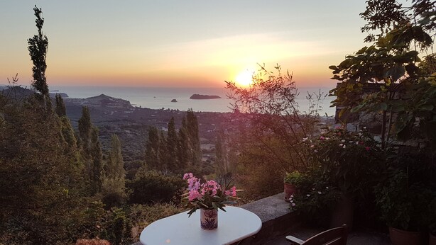 Un patio con una mesa redonda y dos sillas, con vistas panorámicas al mar y las colinas al atardecer.