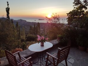 Une terrasse avec une table ronde et deux chaises, offrant une vue panoramique sur la mer et les collines au coucher du soleil.