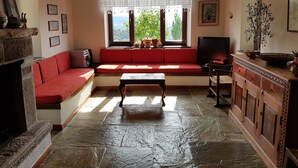 Una sala de estar acogedora con un sofá rojo, suelo de piedra, televisor y ventana con vistas a la vegetación.