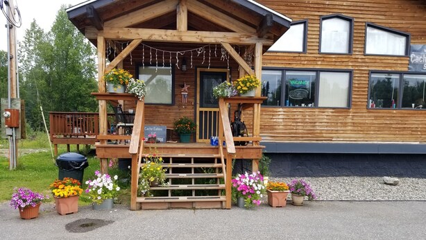 Una cabaña de madera con un porche adornado con flores en macetas, un letrero que indica que es un alquiler vacacional y una vista de un edificio moderno al fondo.