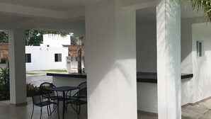 Un espacio al aire libre techado con mesa y sillas, pilares blancos y vista a un edificio al fondo.