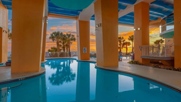 Una piscina interior con vistas al atardecer, rodeada de palmeras y un balcón con barandilla.