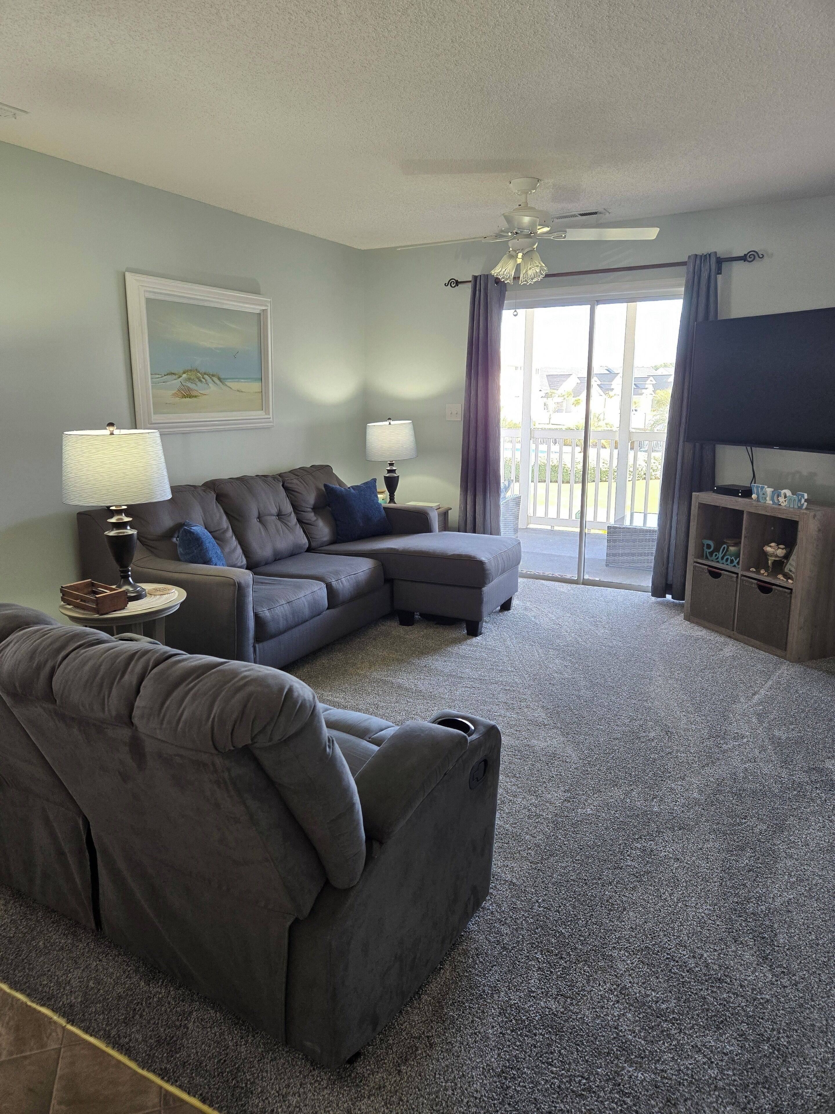 A living room with a sectional sofa, two side tables with lamps, a ceiling fan, a flat-screen TV, and a sliding glass door leading to a balcony.